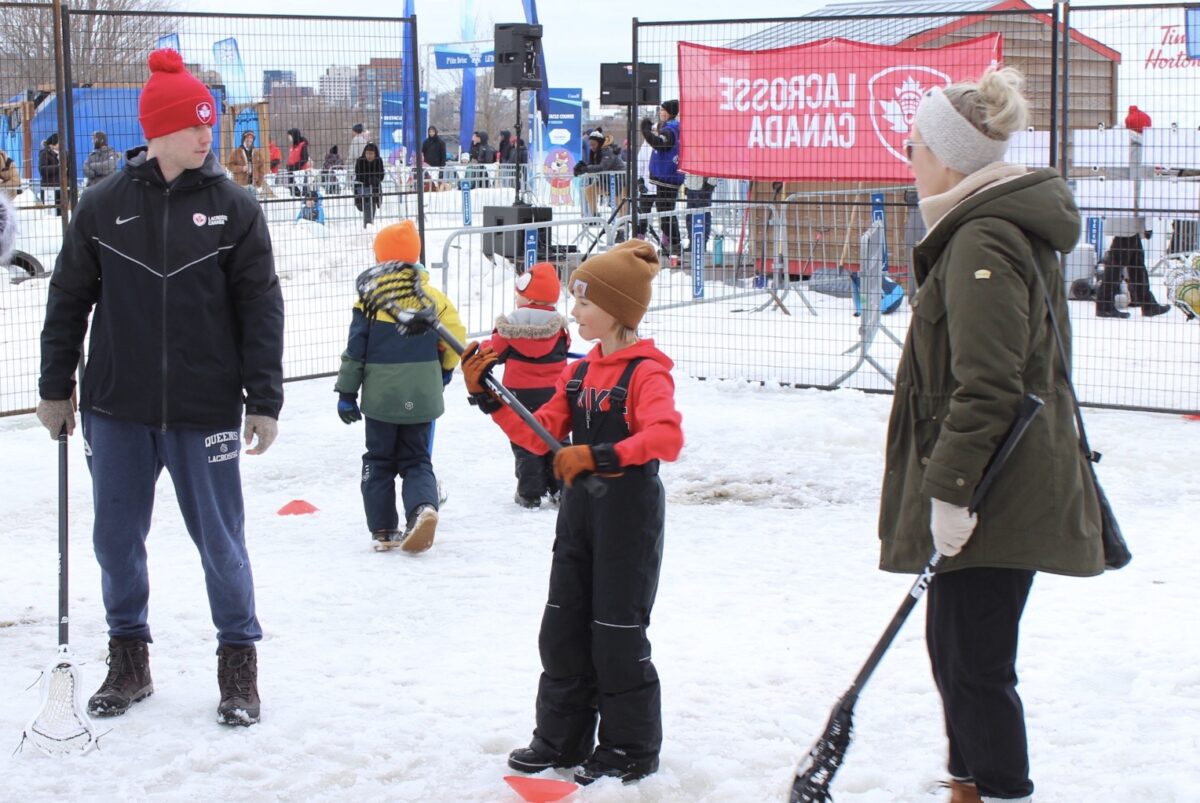 A child holds a lacrosse stick as two adults observe at a snowy activity space