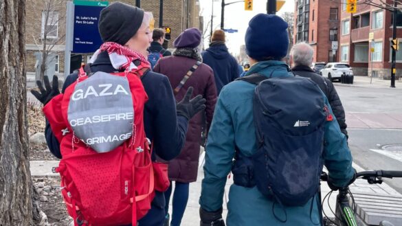 Two protestors walk side by side. The protestor on the left has a red backpack, with a sign tied to it reading "Gaza Ceasefire Pilgrimage." The protestor on the left is walking with a green bicycle.