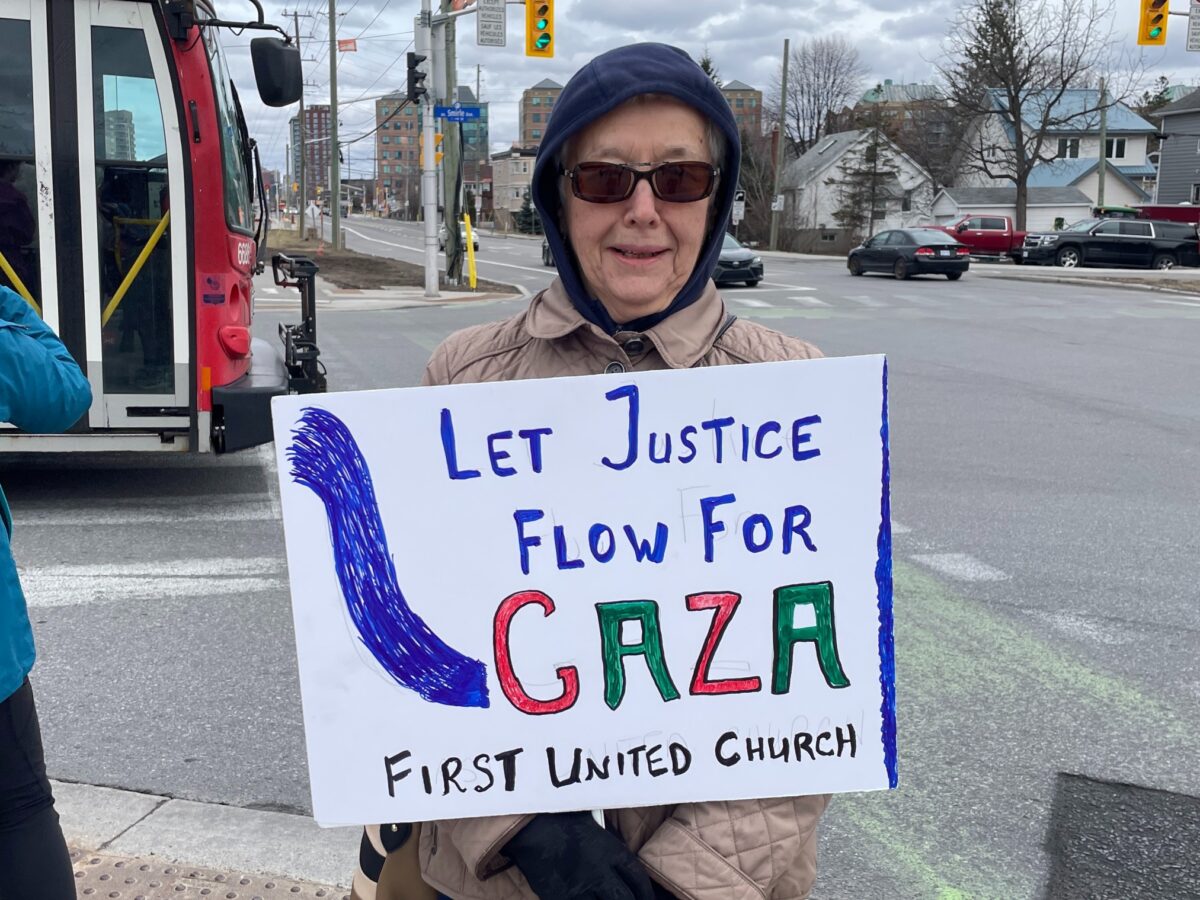 An elderly woman stands on the sidewalk, smiling for the camera. She is holding a hand-drawn sign that reads "Let Justice Flow For GAZA," with the words "First United Church" written in smaller letters below it.