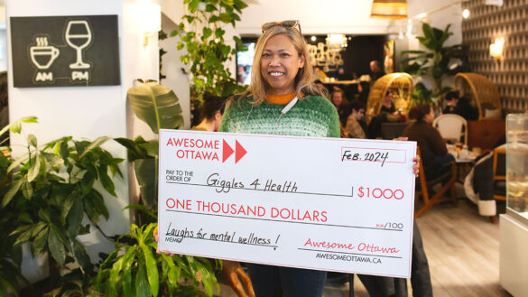 Jocelyn Umengan holds a large check in a cafe from The Ottawa Awesome Foundation for $1,000, celebrating her project Giggles4Health, aimed at fostering community mental wellness and battling loneliness.