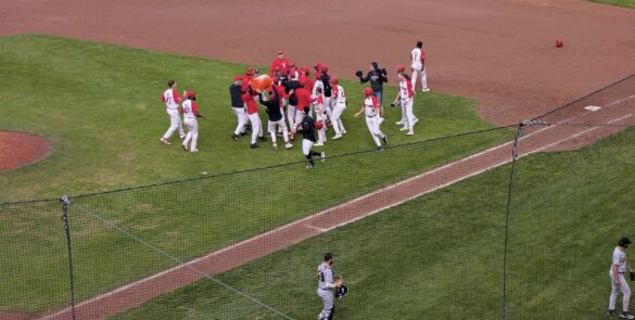 A dozen baseball players converge to celebrate a win between first base and the pitcher's mound at Ottawa Stadium