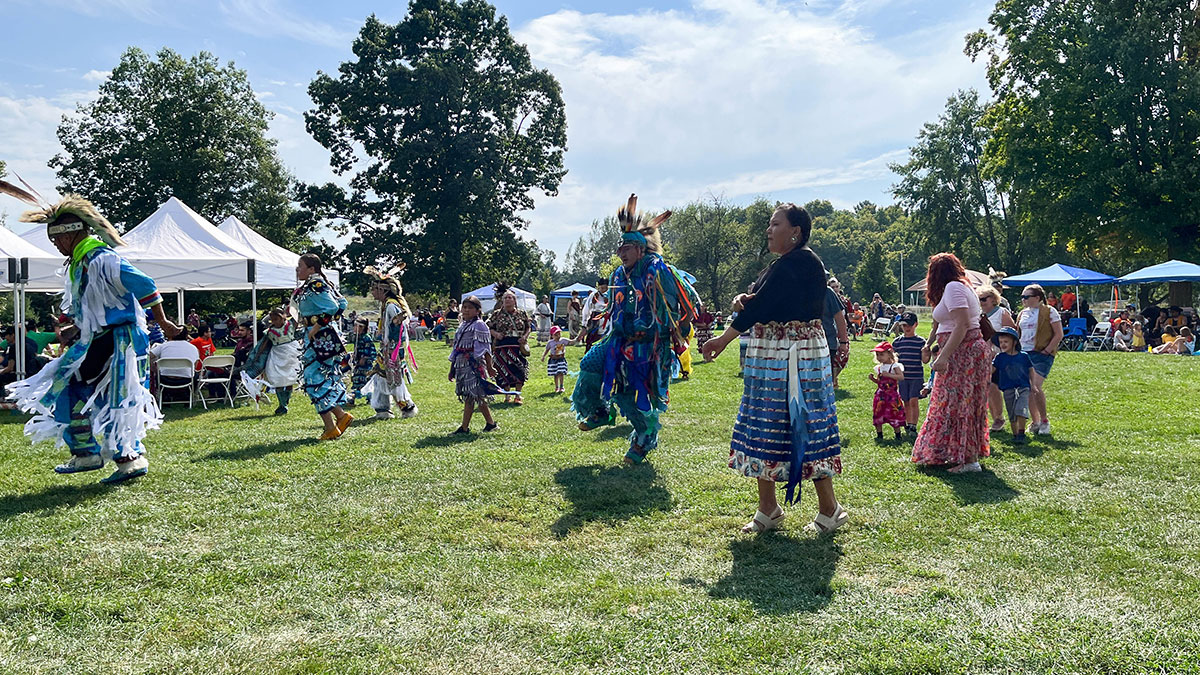 People in brightly coloured regalia dance in park with trees in the background.