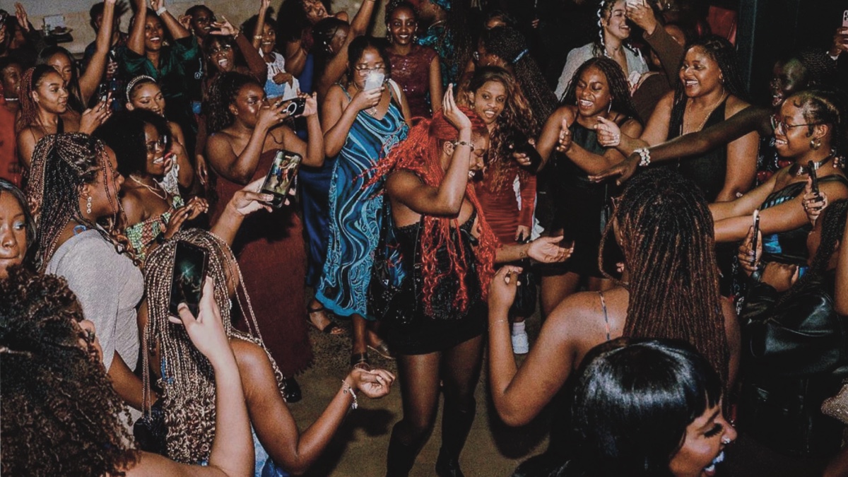 Black women with red braids dancing in the middle of a dance circle with other Black women cheering her on and filming. 