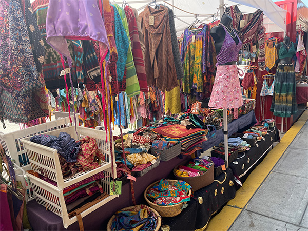 This vendor stall is filled with colourful clothing and accessories. There are tables and racks filled with clothing as well as a few mannequins.