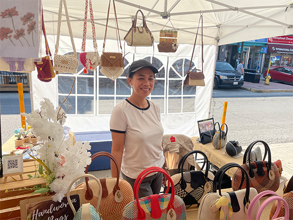 A woman stands under and tent and behind a table covered in handmade bags. There are also bags hanging down from the roof of the tent. She is alone in the stall.