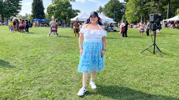 Woman in white top and blue skirt stands in front of circle of people gathered around a white tent in a park.