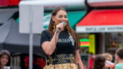 Woman wearing black t-shirt holds a microphone with store fronts in the background.