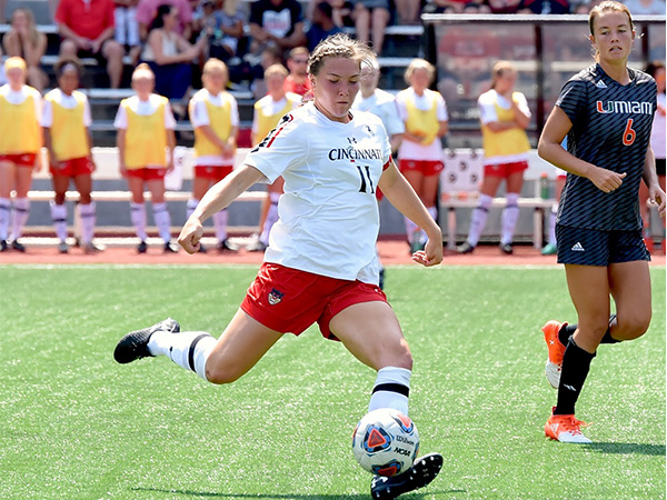 Photo of two women on a soccer field, they are wearing jerseys from opposing teams. The woman on the left has a soccer ball at her feet and is running towards the woman on the right who is defending against her.