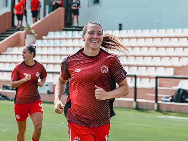 A smiling woman runs towards the camera. She is wearing a red soccer jersey for Team Canada.