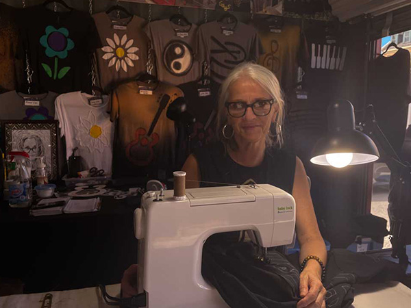 A woman sits behind a table, inside a temporary building. She is at a sewing machine and her handmade shirts are hanging on the wall behind her. 