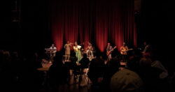 A stage with musical performers is under lights on a stage with red curtains in a darkened performance hall.