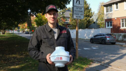 A man holds a fan used for radon services, he's wearing greyish-black shirt with the logo of Budd Radon Services on the right side, with a neighbourhood in the background and a grey car.