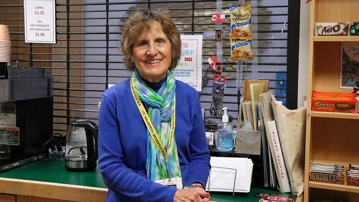 This photo shows a smiling older woman with short brown hair standing behind a counter, likely in a small tuck shop. She is wearing a blue sweater and a colourful green and blue scarf, with a yellow lanyard around her neck that says "LTC Volunteer Bénévole," indicating she is a volunteer. The background includes various items for sale, such as candy bars on a shelf to her right and small snack items like Werther’s Original candies hanging behind her. On the counter, there is a coffee maker, some coffee pots, a bottle of hand sanitizer, and a stack of papers and envelopes. A small sign next to the coffee maker lists prices, with coffee priced at $1.25 and tea or hot apple cider for $1.00. The setting appears welcoming and organized, suggesting a friendly environment.