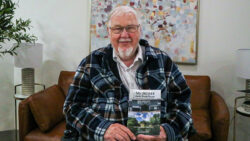 This photo shows a light-skinned, smiling older man with short grey hair holding a book that reads, "My Home Away from Home: My First Year in Long Term Care." He is donning a white inner shirt beneath a green and white striped jacket. The background features a brown cushion and a piece of hanging artwork. 