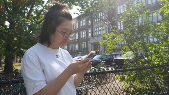 With a school in the background, a grade 12 student goes on her phone. She is wearing a white shirt.