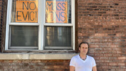 A man in a white shirt stands against a brick wall with one sign in a window behind him that reads "NO MASS EVICTIONS".