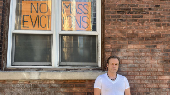 A man in a white shirt stands against a brick wall with one sign in a window behind him that reads "NO MASS EVICTIONS".