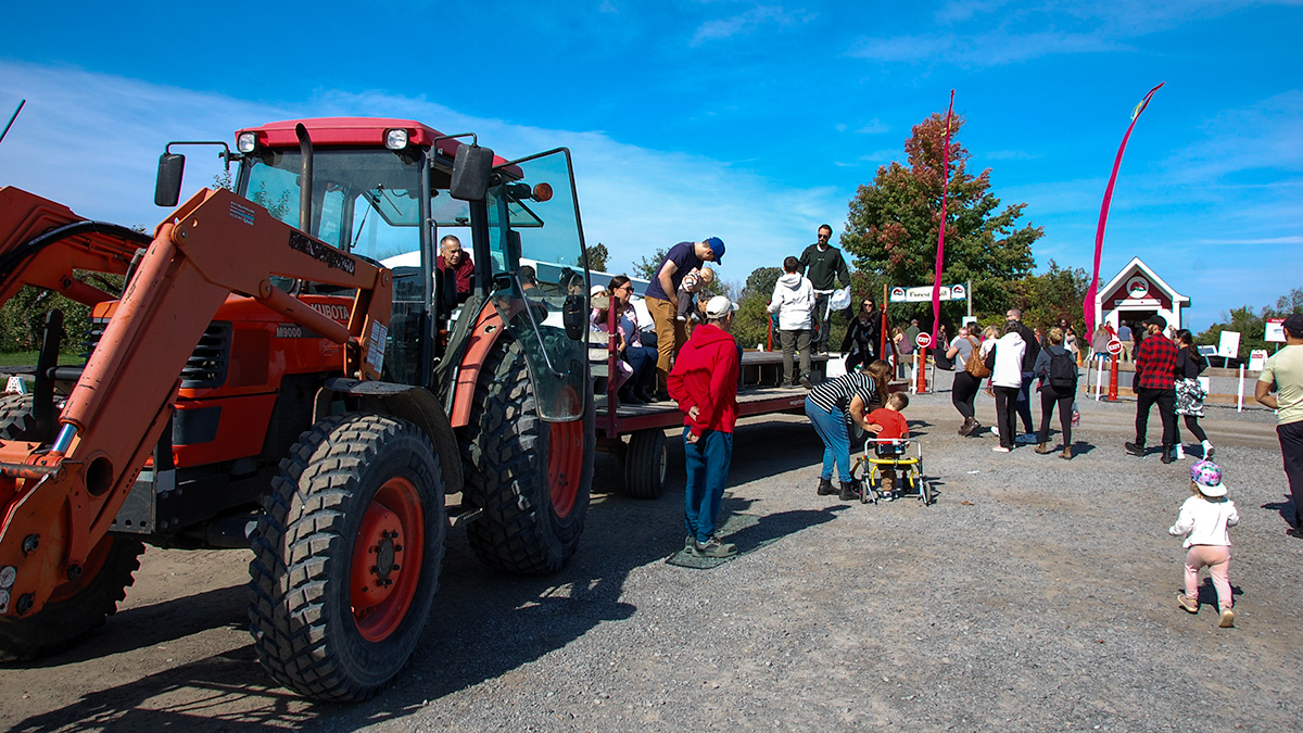 A wagon attached to an orange tractor with adults and children walking to get on board.