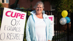 A woman stands in front of a black fence, smiling at the camera. She is wearing a light blue jacket over a white shirt. Behind her, there are signs with the CUPE logo in bold pink letters, one of which reads, "0% equals 0 respect." Three balloons in yellow, blue, and silver are attached to the fence. The scene appears to be part of a labor protest or rally organized by CUPE (Canadian Union of Public Employees).