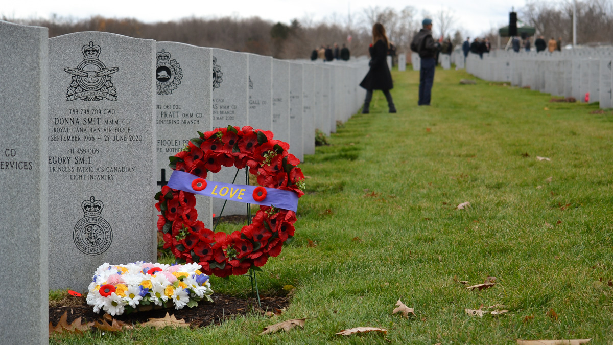 Among tombstones there stands a red poppy wreath with a purple banner across it reading the word love, beside sits a basket of flower and in the foreground there is a man and two flags at half-mast.