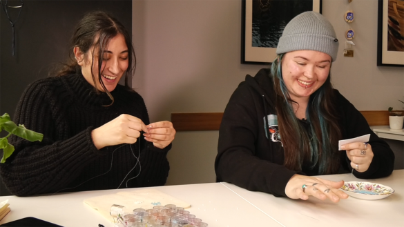 Ely Pittman, on the left, and Jayde Naponse, on the right, are sitting together at a table in a café. In front of them on the table are the beadwork projects they are individually working on,