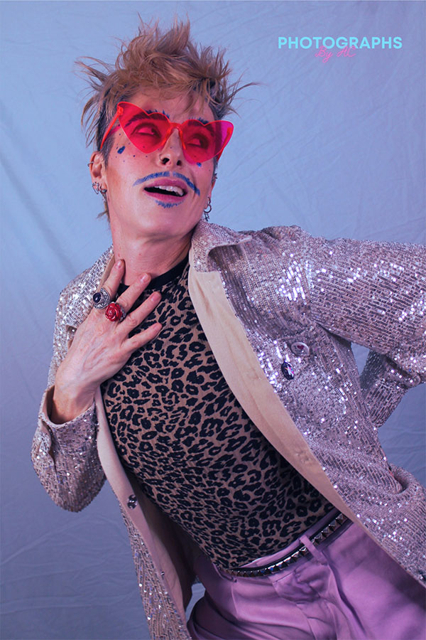 Jax Stellan Fenris Dore is an MX drag king. He is wearing a leopard print shirt, a sparkly pink blazer, purple pants and heart shaped sunglasses. He poses for the picture.