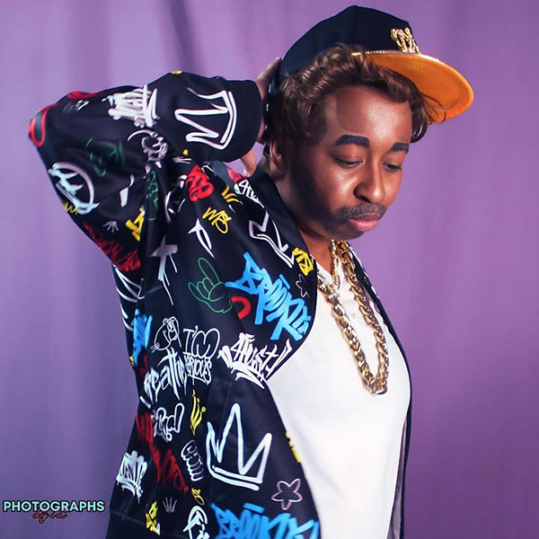 Omari B. Johnson the drag king is wearing a black baseball cap, a gold chain necklace and a graphic bomber jacket. He poses for the picture.