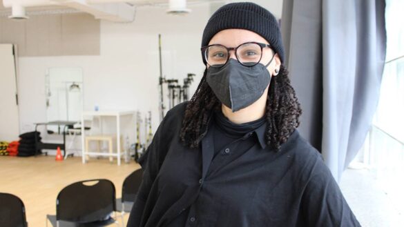 Adrienne Row-Smith is dressed in black, wearing a black mask and hat, and standing in the new studio space with chairs and photography equipment.