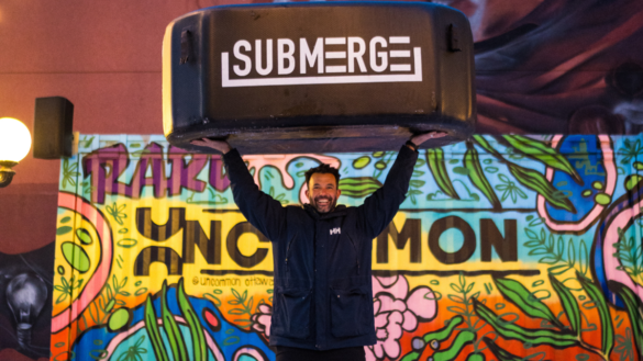 A man, Tarik Hassan, in a winter coat, holding a black cold plunge tub that says Submerge on it right over his head. There is a colourful storage container behind his that spells out Uncommon.