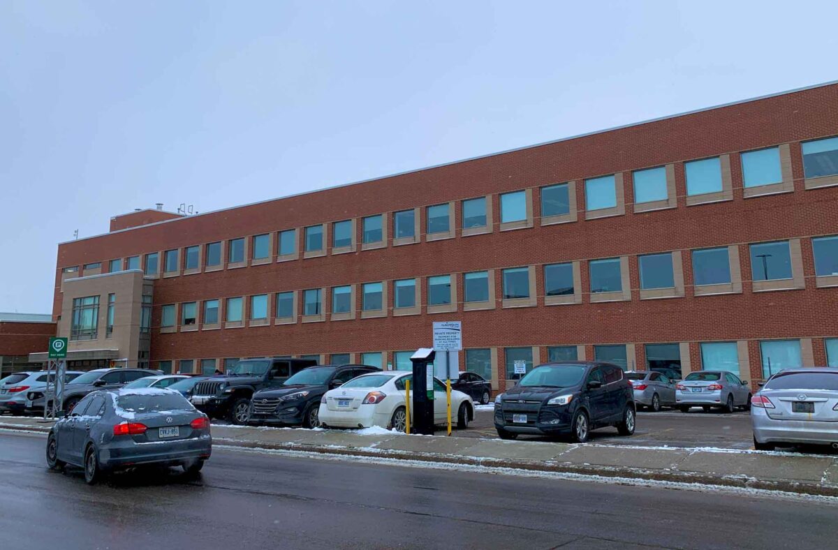 A wide shot photo of the OPS training facility at Algonquin College. A red-bricked building with windows and cars parked in front.