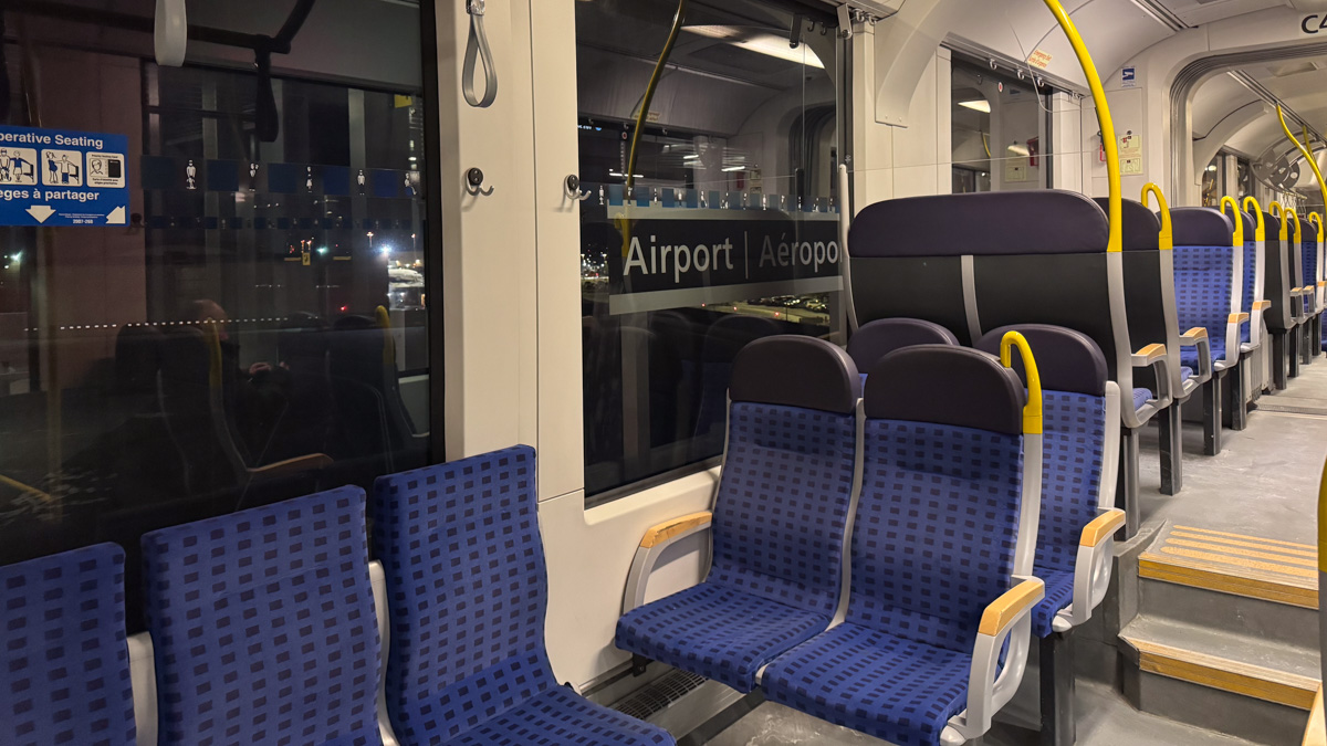 Shot from inside Line 4 of the O-Train. Seats are shown, and the "Airport" sign can be seen through a window.