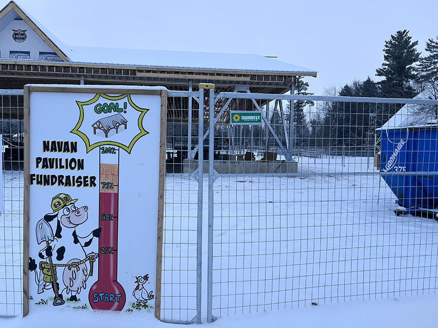 A wooden sign reading "Navan Pavilion Fundraiser" sits on metal gates of a construction site. The sign has a photo of a cow wearing a construction hat and belt, holding a shovel and pointing to a fundraising meter.