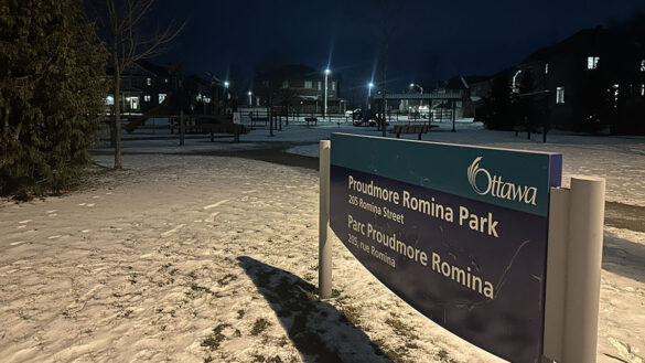 A nighttime photo of the Proudmore Romina park sign slightly to the left, with a view of the parks play structures in the back before rows of residential houses.