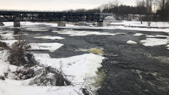 Rideau River's water flowing downstream. A bridge is over the partly iced river.