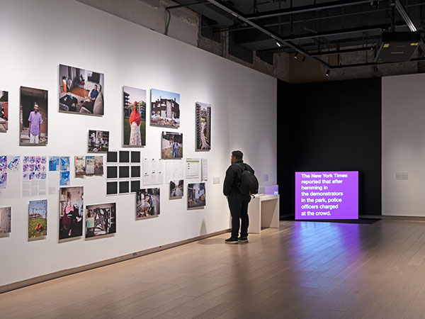 A wall to the left covered in photographs, art prints, and documents with a person standing looking at it. A purple screen to the right reads "The New York Times reported that after hemming in the demonstrators in the park, police officers charged at the crowd."