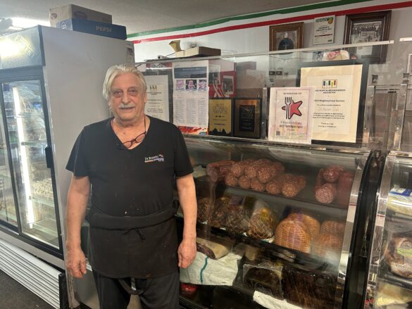 Paolo Di Rienzo stands in front of his deli's meat counter. The smiling grey haired man is wearing a black shirt and black pants whith his glasses hanging from his neck onto his chest