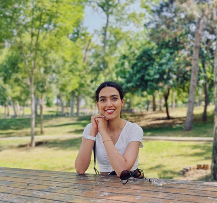 A young woman is seated at a picnic table, smiling, surrounded by greenery