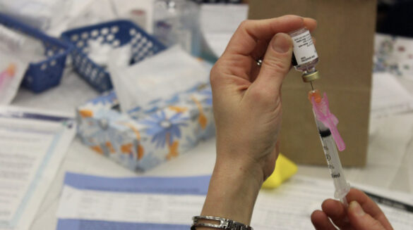 The hands of a doctor or nurse are shown with a syringe extracting vaccine from a small bottle