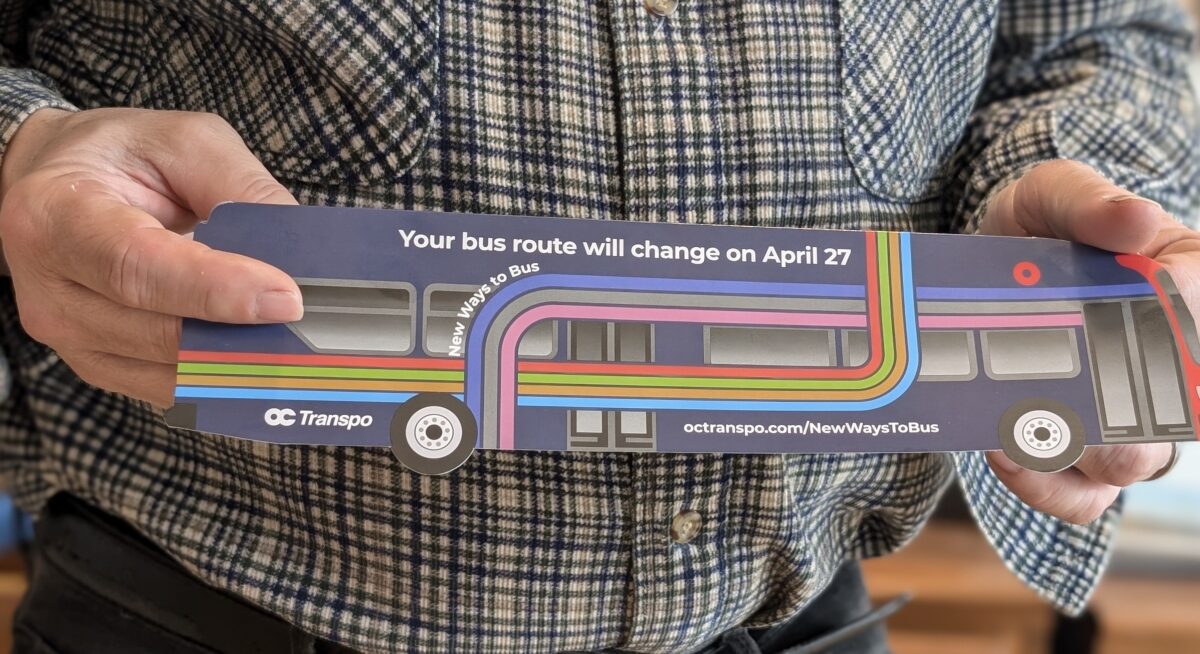 A person holds a foot-long paper bus advertising OC Transpo route changes beginning April 27