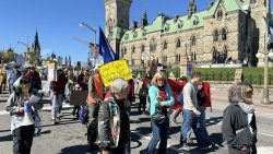 Protesters march down the street, with Parliament Hill behind them, holding a sign that reads "Fossil fools- Ruining our children's future."