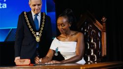 Solange Tuyishime Keita wearing a white gown and her Order of Ottawa medal, signs a document while Mayor Mark Sutcliffe, in his ceremonial chain of office, stands beside her.