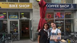 two people stand in front of a building with a sign that says, "Bike shop" and "Coffee"