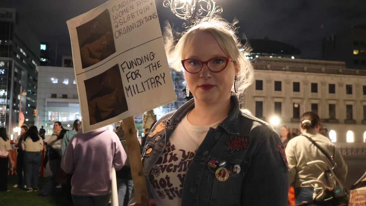Julie Lalonde stands with a sign referring to the government's lack of funding towards women and 2SLGBTQIA+ organizations.