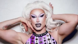 A photo of drag queen Sunshine Glitterchild, who is known as Eric Vance out of drag. Sunshine is wearing a voluminous blonde wig. Her arms are raised, bent at the elbows and stretching inwards towards her face, making a 'vogue'-like pose. The top part of a purple and white vertically striped dress she's wearing is visible.