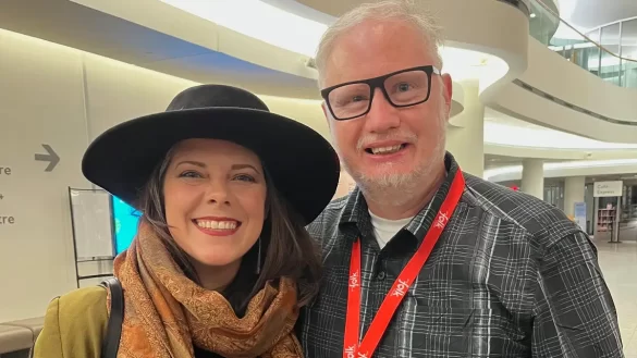 Melanie Brulée and Martin Noakes shoulder to shoulder at the 2025 Canadian Folk Music Awards. Brûlée wears a large brimmed black hat (her signature). They both smile ear to ear with their red passes for the event at the Canadian Museum of History in Gatineau Qc. [Photo courtesy of Great Dark Wonder]