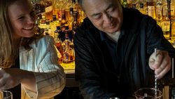 Female Mixologist laughs at Peter Hum stirring a drink. The background shows a bar full of alcohol bottles.